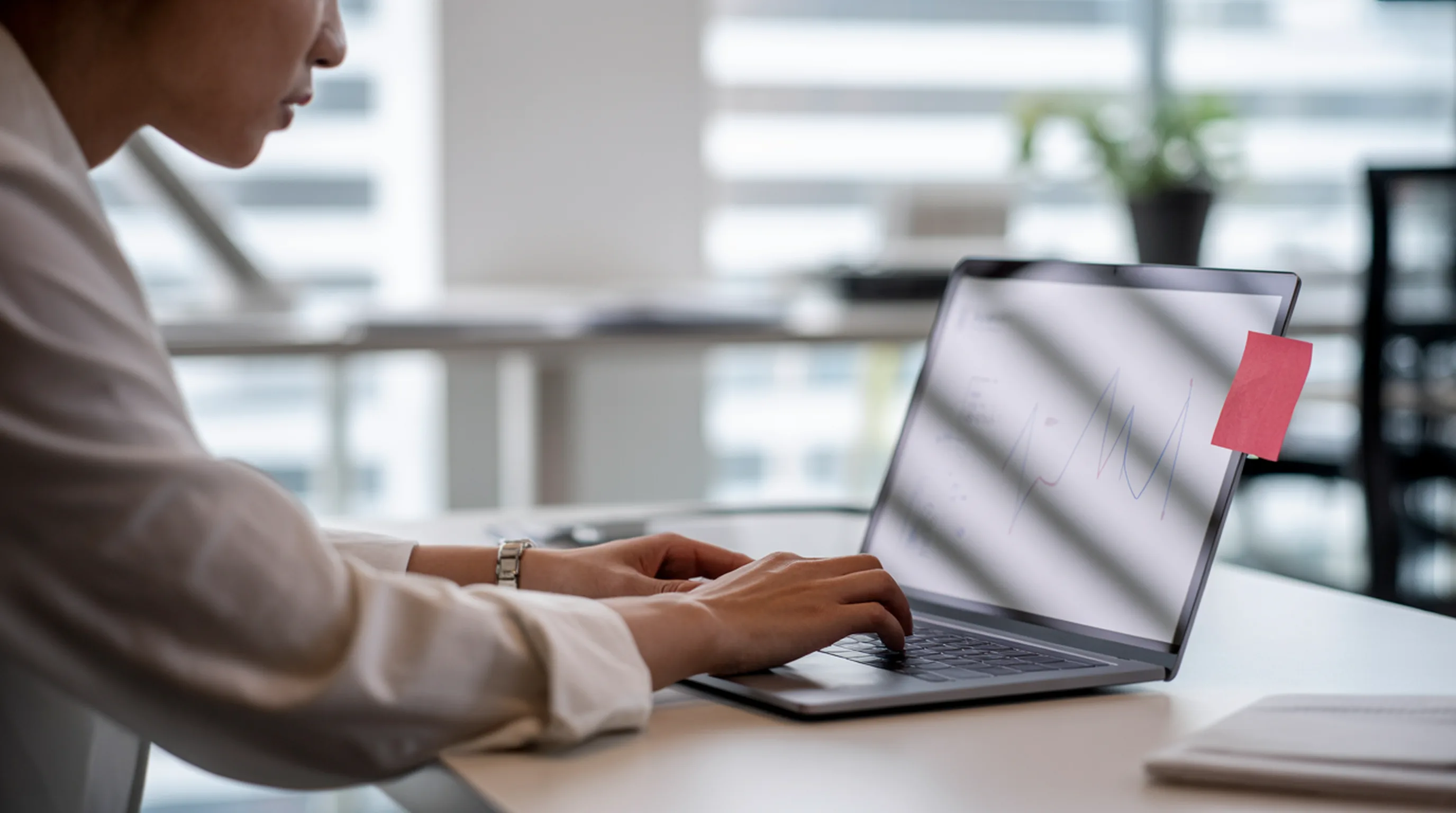 A professional typing on a laptop at an office desk, the screen showing a soft line chart and a red sticky note on its edge.
