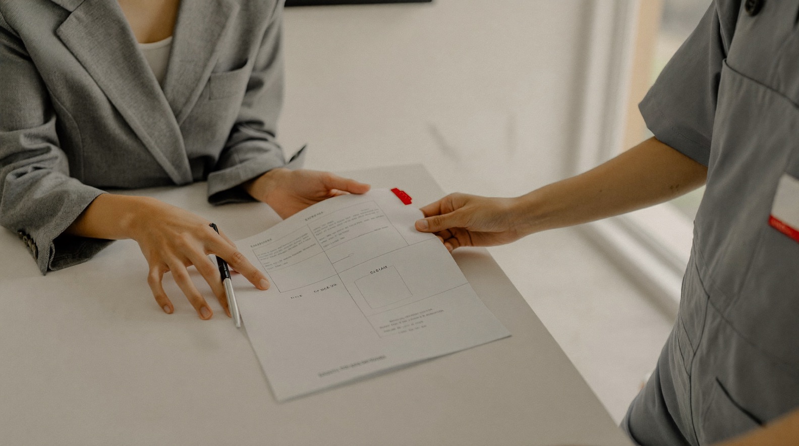 Two people at a table, one in a blazer and one in work coveralls, reviewing a printed proposal document together with a red tab bookmark on one page. An illustration of the proposal handover described in stage 03.