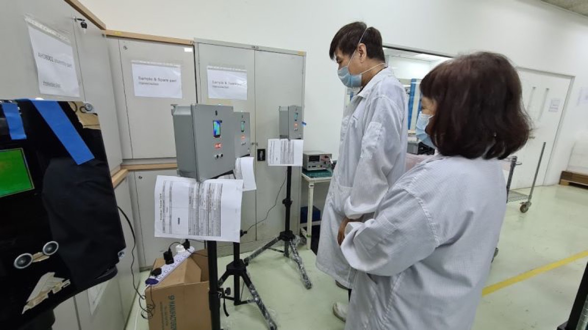 Two staff in cleanroom coats at the quality control station, with thermal imaging units mounted on tripods and printed test records posted beside them.