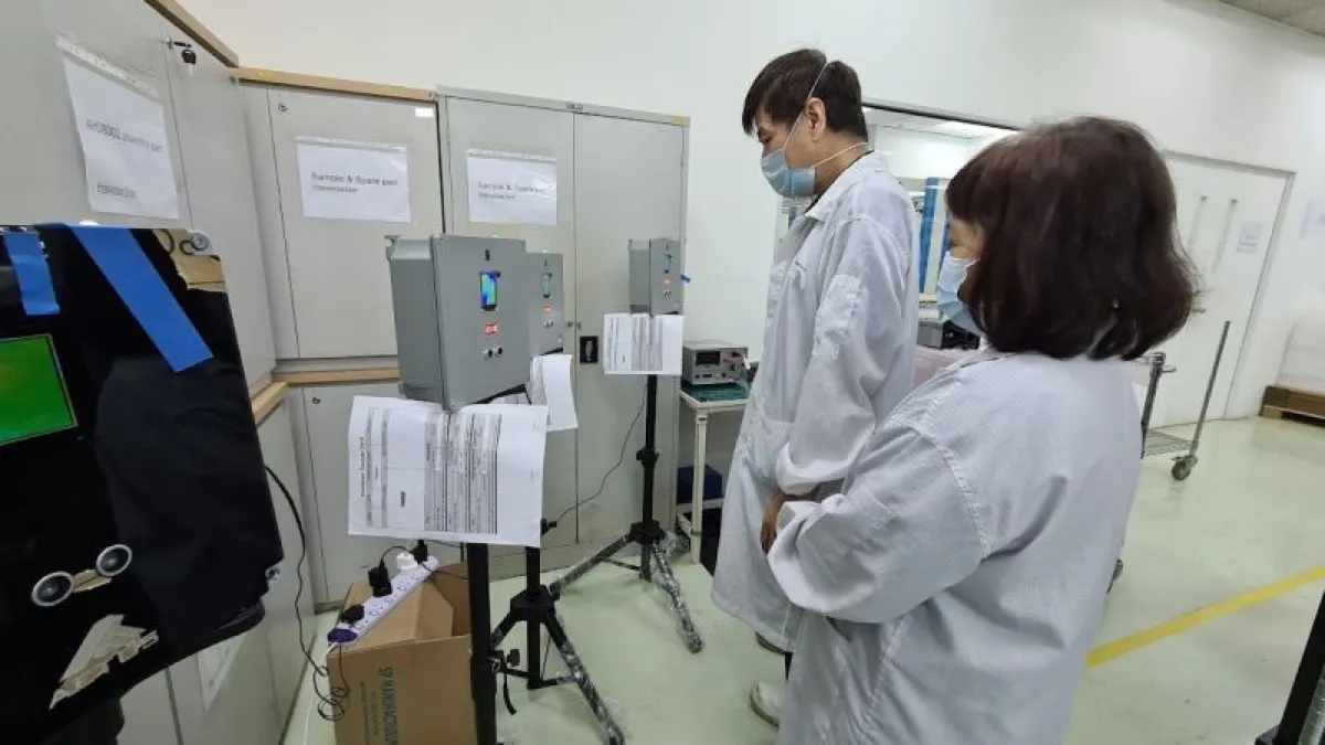 The AETOS KoolScan quality control station, with two staff in cleanroom coats, thermal imaging units mounted on tripods, and printed test records posted beside them.