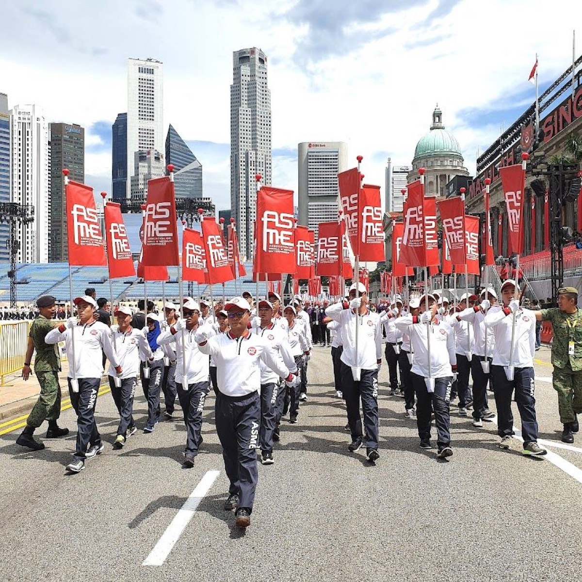 The ASME marching contingent on the parade route, banners raised, with the city skyline behind.