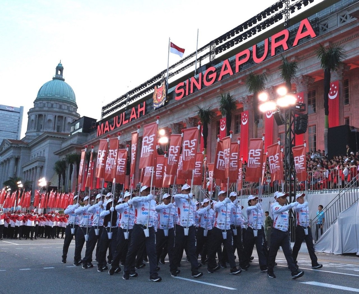 The Association of Small and Medium Enterprises contingent marching at the National Day Parade 2019, red flags raised, in front of City Hall lit up for the parade.