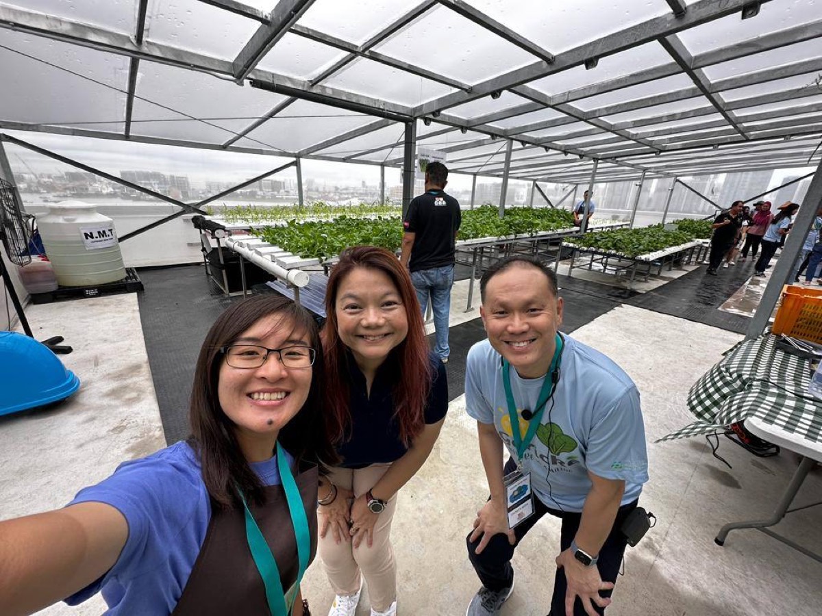 Three members of the project team at the rooftop farm during the open house, with planted growing channels and visitors behind them.